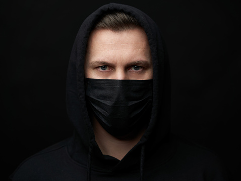 Young man wearing face mask. Handsome man in black hoodie wear black medical mask, gray background, copy space. Pandemic coronavirus covid-19 quarantine period concept.