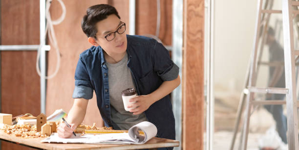 asian attractive glasses interior designer working at house site renovation construction progress with smile and confident at desk with blue print and drawing tool