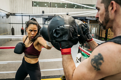 Hardworking Hispanic female boxer in early 20s developing effective punching technique while working out with focus mitts held up by male trainer.