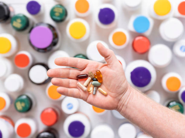 many nutritional health supplements and vitamins in capsules and tablets in a hand before consuming them, with many colorful food supplements and vitamin bottles out of focus in the background on a white table, shot directly above with focus on the hand. - vibrant color healthcare and medicine healthy lifestyle vitamin pill imagens e fotografias de stock