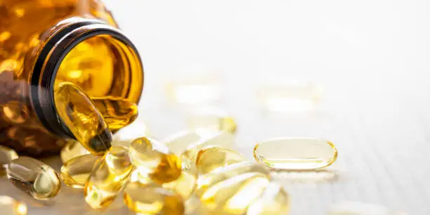 Many nutritional health supplement fish oil capsules spilling out of their bottle onto a white wood table background, shallow depth of focus. Very shallow depth of field with focus being on the rim of the bottle and one isolated capsule on the right of the image. Good copy space.