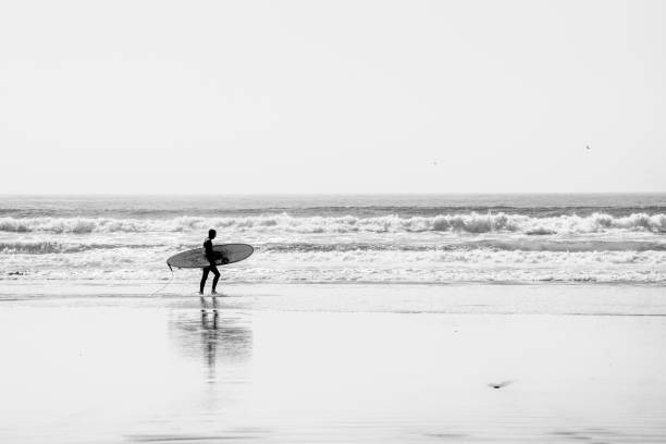Atlantic Ocean at Taghazout, a tourist area in Morocco that is receiving considerable investment in new hotels, apartments etc. In the foreground is a surfer with a surf board walking into the surf across the wet sand.