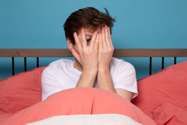 istock man sitting on bed peeping through fingers, frightened 1215685174