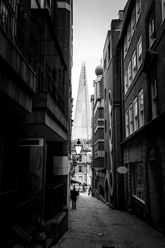 Monochrome image depicting people walking on a narrow cobblestone London street flanked by old fashioned architecture. In the distance the ultra modern, futuristic structure of The Shard (the tallest building in wester Europe) is an imposing presence.