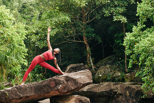 Pretty fit Black woman doing triangle pose on big rock in the forest