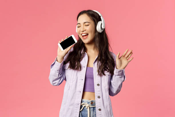 retrato de cintura arriba de la mujer feliz con estilo utilizando auriculares para escuchar nueva pista impresionante, descargar aplicación de karaoke y el uso de teléfono inteligente como micrófono, cantando a lo largo de la música impresionante - waistup fotografías e imágenes de stock