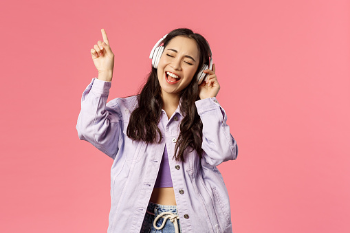 Portrait of attractive carefree asian girl chilling and vibing, listening music in headphones, raising finger up while trying sing along and reach highest note in song, pink background.