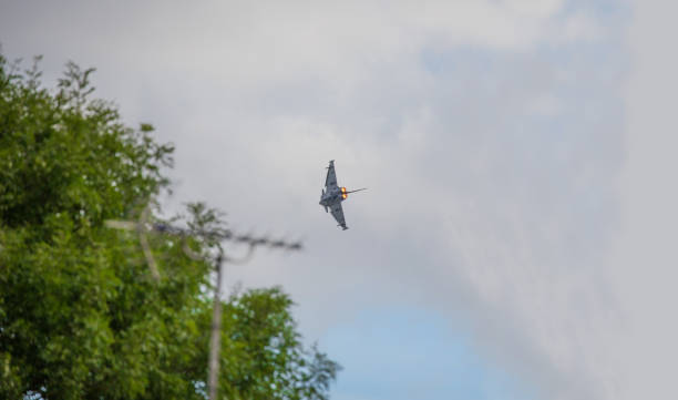 eurofighter "typhoon" z pełnymi dopalaczami latającymi nad bazą fairford air force w gloucestershire. telewizor mieszkalny, antena i drzewa na pierwszym planie, podkreślające wpływ lotnictwa na środowisko - military airplane military eurofighter typhoon zdjęcia i obrazy z banku zdjęć