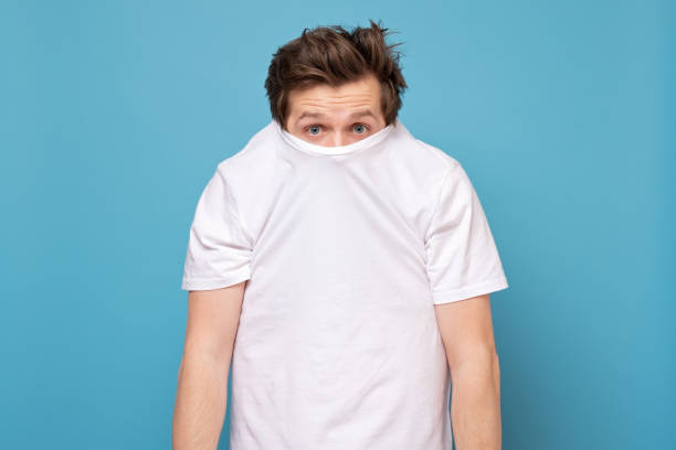 Shy caucasian man hiding behind his white shirt being afraid of coronavirus. Shy caucasian man hiding behind his white shirt being afraid of coronavirus. Studio shot on blue wall. shy stock pictures, royalty-free photos & images