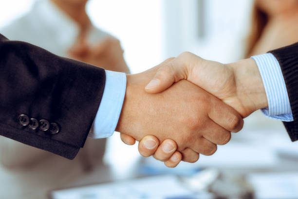 istock Business people shaking hands at meeting while theirs colleagues clapping and applauding. Group of unknown businessmen and women in modern white office. Success teamwork, partnership and handshake concept 1215693544