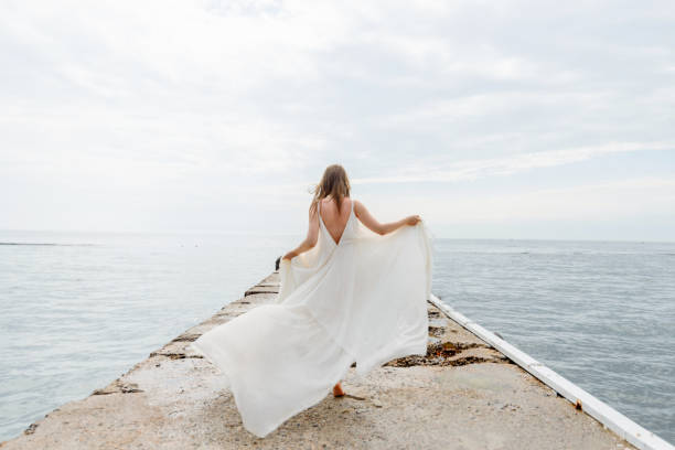 A young beautiful girl in a long milk-colored dress walks along the beach and pier against the background of the sea. stock photo