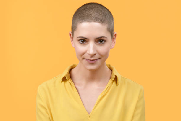 verticale de studio d’une belle jeune femme caucasienne avec la tête rasée sur le fond jaune lumineux. portrait de survivant de cancer. - shaved head photos et images de collection