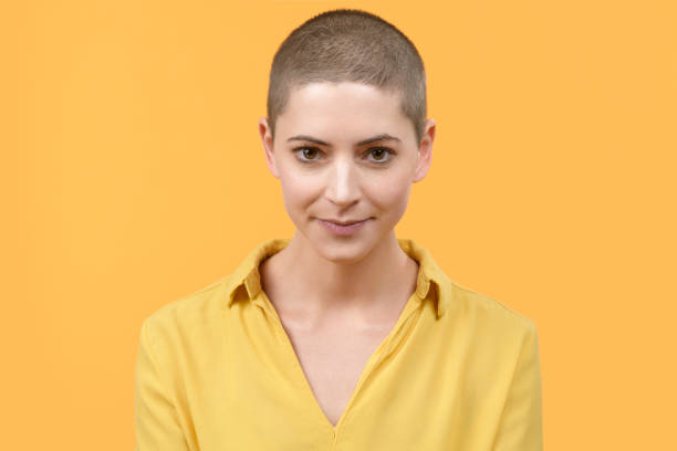 ritratto in studio di una bella giovane donna caucasica con la testa rasata su sfondo giallo brillante. ritratto sopravvissuto al cancro. - shaved head foto e immagini stock