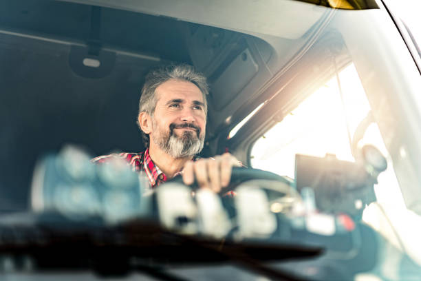 Happy mature truck driver driving his truck on a sunny day. Front view of truck driver sitting in his truck and driving to a warehouse. driver occupation stock pictures, royalty-free photos & images