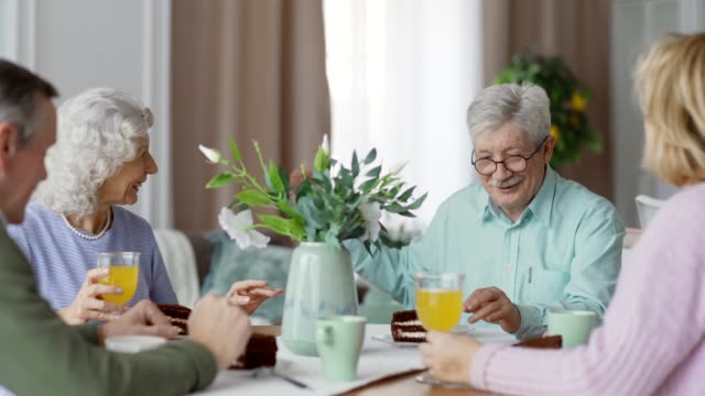 Group of four senior friends sitting at dining table at home, talking cheerfully over dessert and laughing