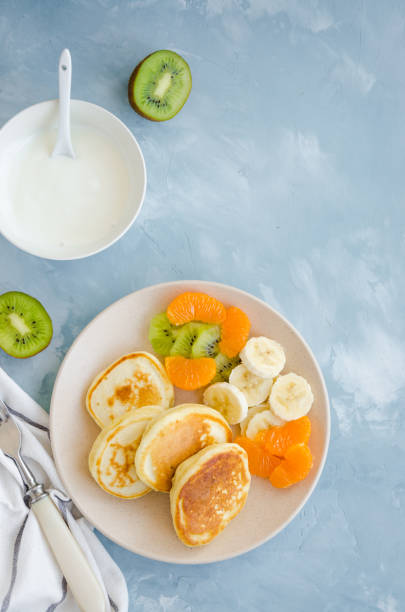 istock Pancakes on buttermilk or kefir with fresh fruit and yogurt on a plate on a light blue concrete background. A delicious and healthy breakfast. Vertical orientation. Top view, copy space. 1215707578