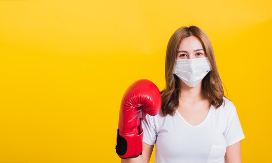 Portrait Asian Thai beautiful happy young woman wearing mask protection and boxing gloves her get ready for Coronavirus fighting against, studio shot isolated on yellow background, with copy space