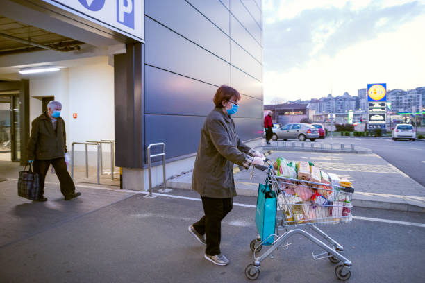 Old woman with protective medical mask and gloves leaving grocery shopping store with shopping cart full of groceries. Panic buying during coronavirus senior shopping hours. Coronavirus lockdown. stock photo