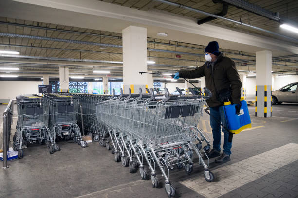 Grocery shopping store worker with face protection mask and protective gloves spraying shopping carts using chemical agents to stop spreading virus infections. Disinfection for virus killing inside shopping store garage. stock photo