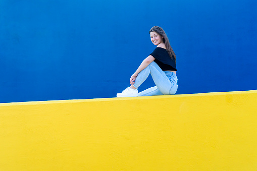 Young beautiful woman with long hair sitting on yellow wall while looking happy to camera in a sunny day