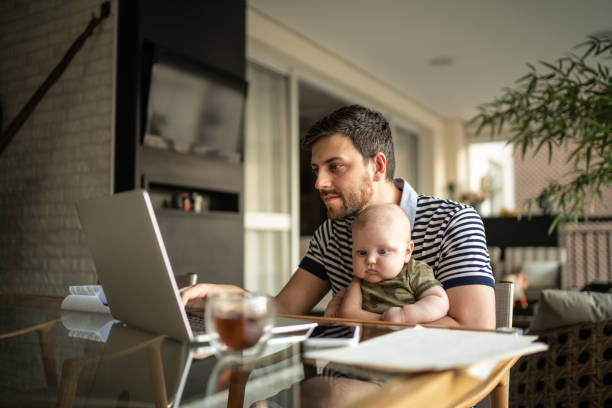 homme retenant son fils nouveau-né et travaillant avec l’ordinateur portatif à la maison - baby carrier photos photos et images de collection