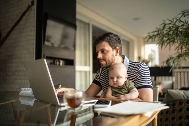 man holding his newborn baby son and working with laptop at home - baby carrier fotos imagens e fotografias de stock