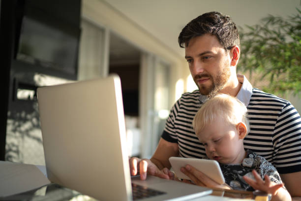 istock Man holding his son and working with laptop at home 1215722757
