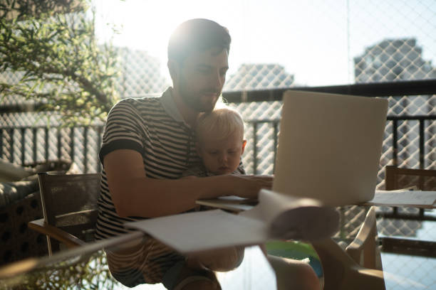 istock Man holding his son and working with laptop at home 1215723073