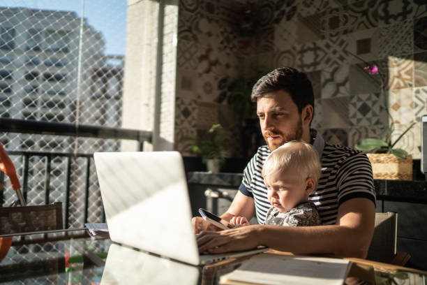 istock Man holding his son and working with laptop at home 1215724684