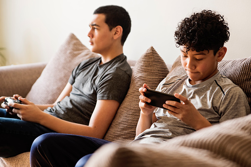 Teenage boys entertaining themselves in small city apartment self-isolating from Covid-19. They are latin american, one is playing video games the other is playing with mobile device. Horizontal waist up indoors shot with copy space.