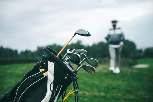 Close-up shot of a golf bag in a golf course. There is an unrecognizable defocused person in background, focus on the golf bag. Horizontal shot.