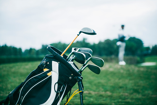 Close-up shot of a golf bag in a golf course. There is an unrecognizable defocused person in background, focus on the golf bag. Horizontal shot.