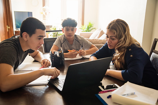 Mother homeschooling teenage boys from home on dinner table in small city apartment self-isolating from Covid-19. Mother and boys are latin american. Horizontal waist up indoors shot with copy space.