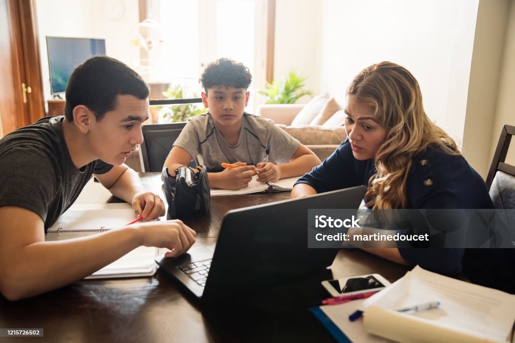 Mother homeschooling teenage boys in self-isolation, Covid-19. Mother homeschooling teenage boys from home on dinner table in small city apartment self-isolating from Covid-19. Mother and boys are latin american. Horizontal waist up indoors shot with copy space. Teenager Stock Photo
