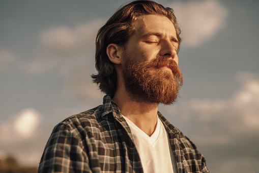 From below side view of tranquil peaceful adult bearded male in checkered shirt standing with eyes closed against cloudy sky in sunny day