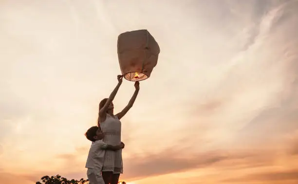 From below side view of little boy embracing teen sister while girl releasing sky lantern and together making wish, standing against sunset sky in summer evening