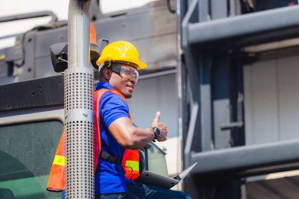 istock Worker in hardhat and safety vest holding laptop standing on container stackers control loading containers box from cargo, Smiling man and giving thumbs up 1215735931