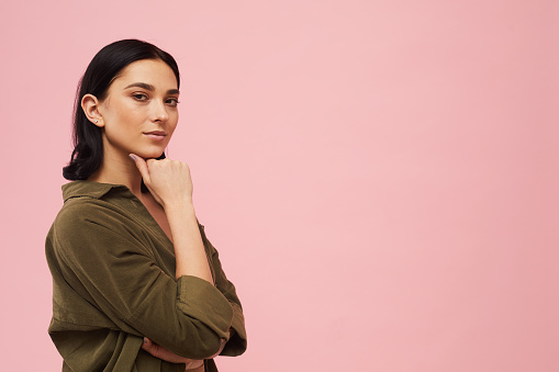 Waist up portrait of modern young woman posing confidently and looking at camera while standing against pink background in studio, copy space