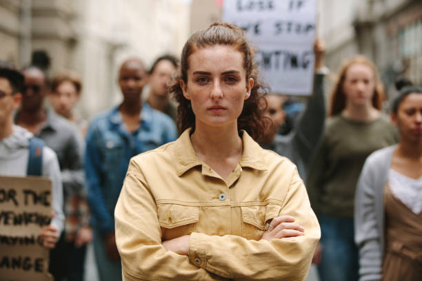 Female activist leading a protest Young woman standing with arms crossed standing in a rally. Woman protesting with group of activists outdoors on road. activist stock pictures, royalty-free photos & images