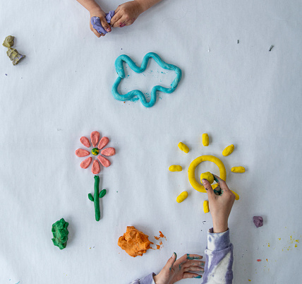 An aerial view of two young children play together with modeling clay and play-dough.  A flower, could and sunshine can be seen n the middle of the photo that have already been created, and they continue to mold and create.