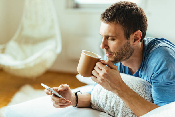 молодой человек пьет кофе во время использования мобильного телефона в спальне. - drinking tea cup drink стоковые фото и изображения