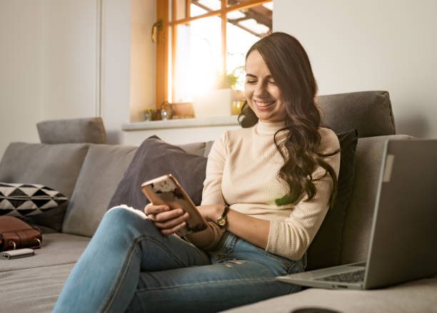istock Smiling young woman using smart phone and laptop on sofa 1215760309