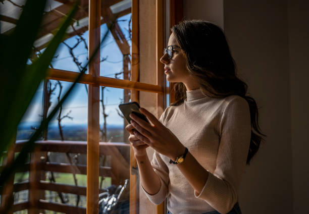 istock Young woman with smart phone looking out window 1215760348
