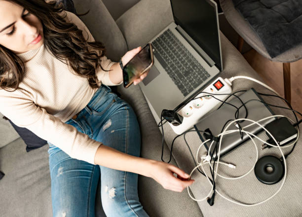 mujer joven cargando dispositivos tecnológicos en el sofá - plug adapter charging mobile phone battery charger fotografías e imágenes de stock