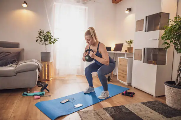 Young woman watching smart phone and squatting with kettlebell on yoga mat in living room