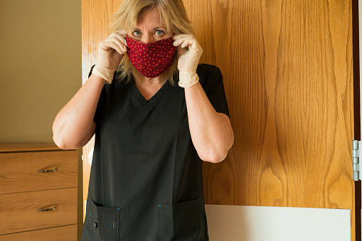 Blond female healthcare worker adjusts her fabric face mask in a patient's room during the corona virus COVID-19 pandemic during a shortage of medical face masks