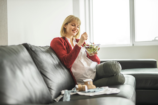 Beautiful pregnant woman relaxing at home, eating salad, getting ready for the new family member arrival, folding baby clothes
