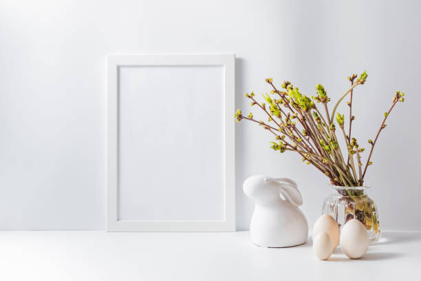 istock Home interior with easter decor. Mockup with a white frame and green buds on branches in a vase on a light background 1215773382
