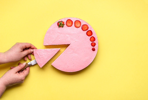 Woman slicing a strawberry cheesecake slice, on a yellow seamless background. Serving a no-bake cheese cake with red berries. Summer fruity pie.