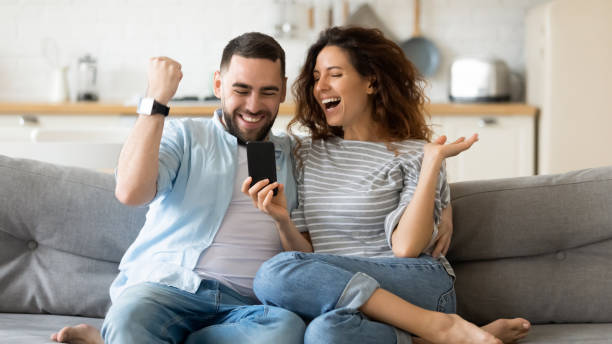 Excited young couple triumph reading news on cell Excited millennial husband and wife relax on couch in kitchen feel euphoric win lottery online on cellphone, overjoyed young couple triumph celebrate read good unexpected news on smartphone gadget unusual couple stock pictures, royalty-free photos & images